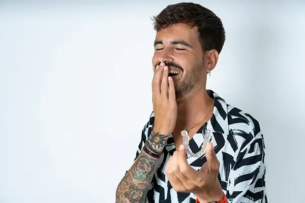 Tattooed man laughing while holding a clear aligner, showcasing its ease of use for adults seeking straighter teeth.